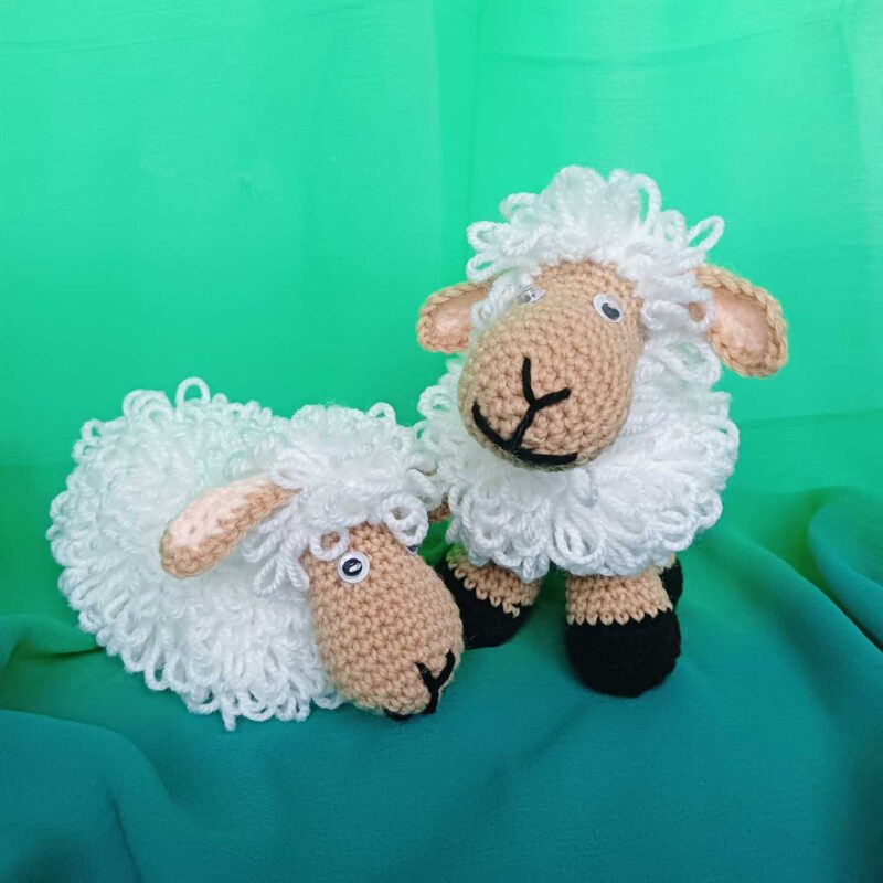 Na zielonym materiale dwie owieczki zrobione ręcznie na szydełku ściegiem pętelkowym. Jedna owieczka stoi na nogach, a druga obok w pozycju leżącej bez widocznych nóg.