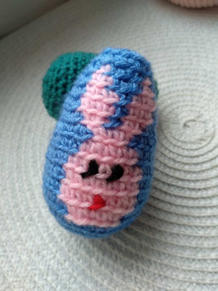 Jajko z niebieskiej włóczki zrobione na szydełku z obrazkiem głowy królika w kolorze różowym leżące na podkładce.