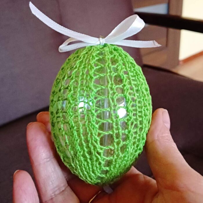Dłoń trzymająca jajko plastikowe o średnicy 8 cm ubrane w koszulkę wykonaną z białej nici na drutach, zawiązana na jajku zieloną kokardką