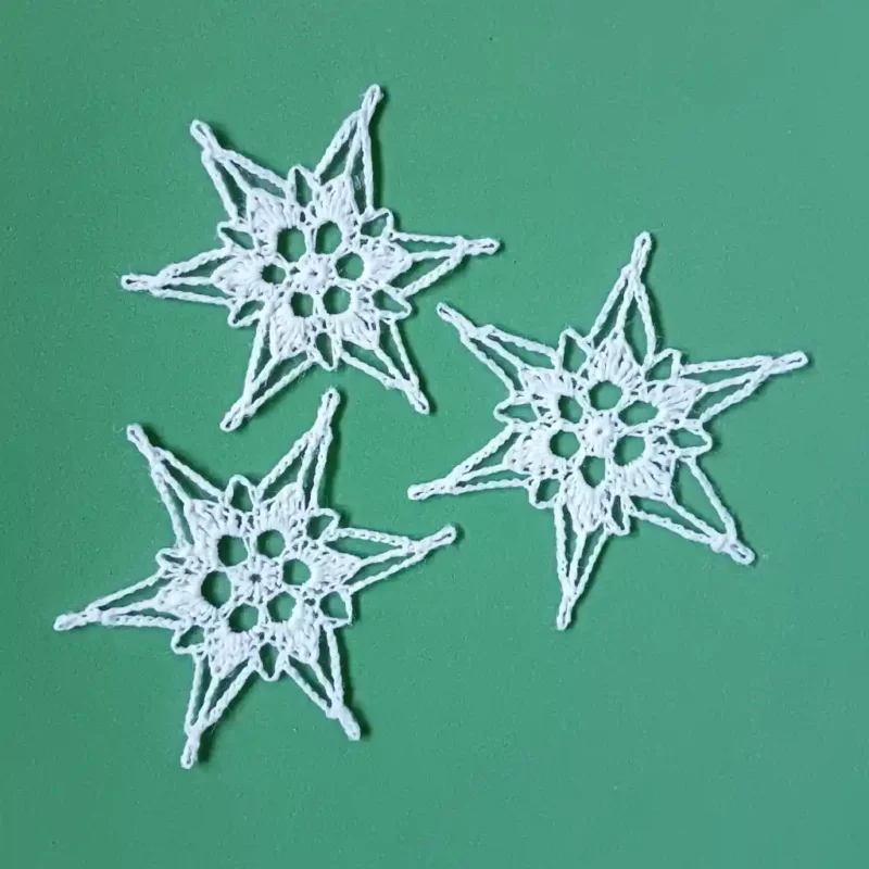 Trzy gwiazdki choinkowe zrobione na szydełku z białego kordonka leżące na zielonym tle