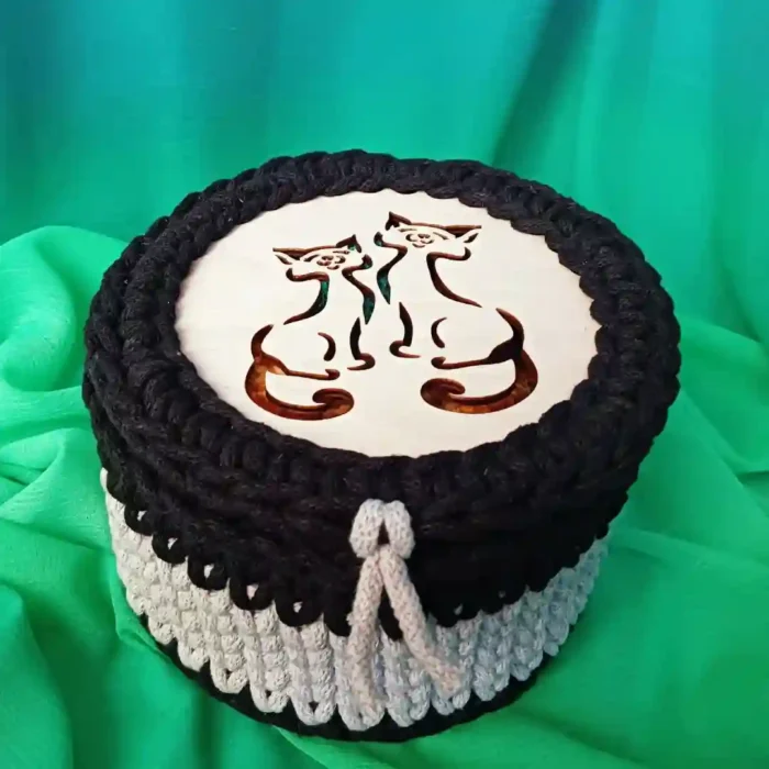 Szaro-czarny Koszyczek-szkatułka ze sznurka bawełnianego z ozdobną jasną pętelką i drewnianą pokrywką, na której znajduje się wycięty laserowo motyw dwóch kotów, zaprezentowany na pofałdowanym zielono-niebieskim materiale.