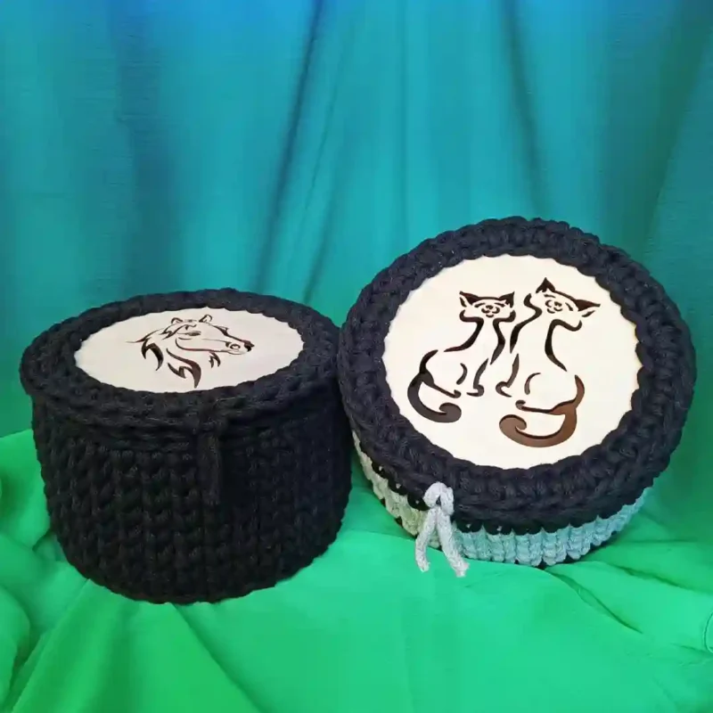 Koszyczek- szkatułka ze sznurka_6 Koszyczek-szkatułka ze sznurka bawełnianego. Ręcznie robione, 2 szydełkowane pudełka na pamiątki z przykrywkami, stojące obok siebie na pofałdowanym zielonym i niebieskim materiale. Pudełko po lewej jest mniejsze, wykonane z czarnego, grubego sznurka, a na jasnej wstawce pokrywki znajduje się czarna grafika profilu głowy konia. Pudełko po prawej jest większe, ze sznurka w czarno-białe paski, a jasna wstawka pokrywki przedstawia grafikę dwóch siedzących kotów z ogonami tworzącymi serce; z boku pokrywki wystaje mała biała sznurkowa pętla.