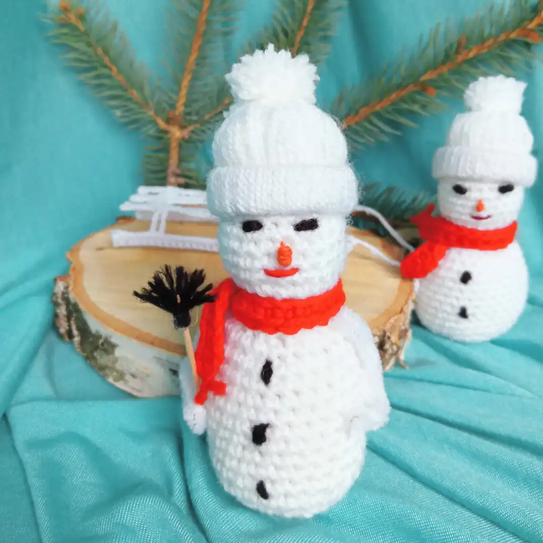 Na pierwszym planie bałwanek zrobiony na szydełku z białej włóczki, w tle plaster drewna, saneczki i jeszcze jeden bałwanek.