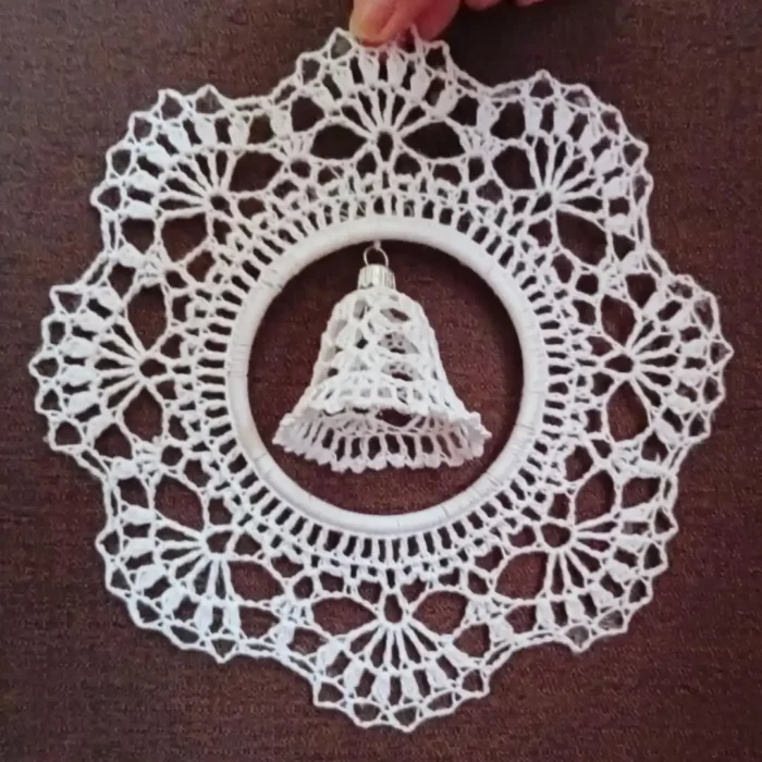 Zdjęcie przedstawia dłoń trzymającą zawieszkę z dzwoneczkiem zrobioną na szydełku. W tle brązowa tkanina.