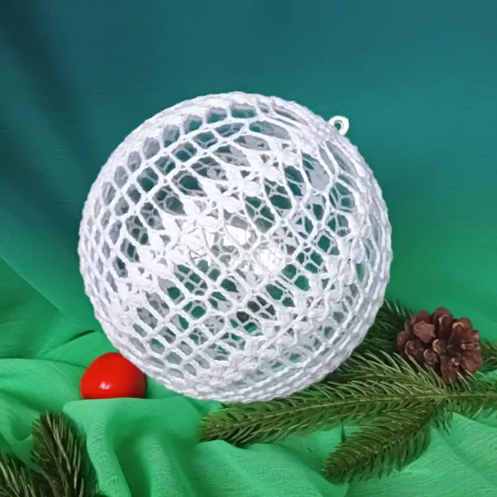 Bombka 15 cm zrobiona na szydełku leżąca na zielonym materiale, obok bombki ułożone gałązki świerkowe, szyszka i jabłuszko.