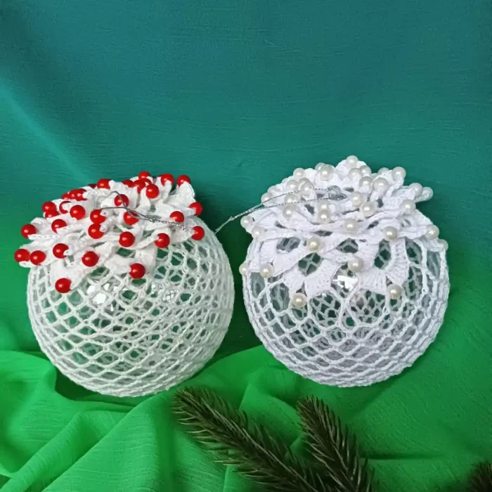 Na zdjęciu 2 bombki szydełkowe zrobione z wzorem kwiata z koralikami. W jednej bombce są czerwone koraliki, a w drugiej perłowe. Tło stanowi zielony material.