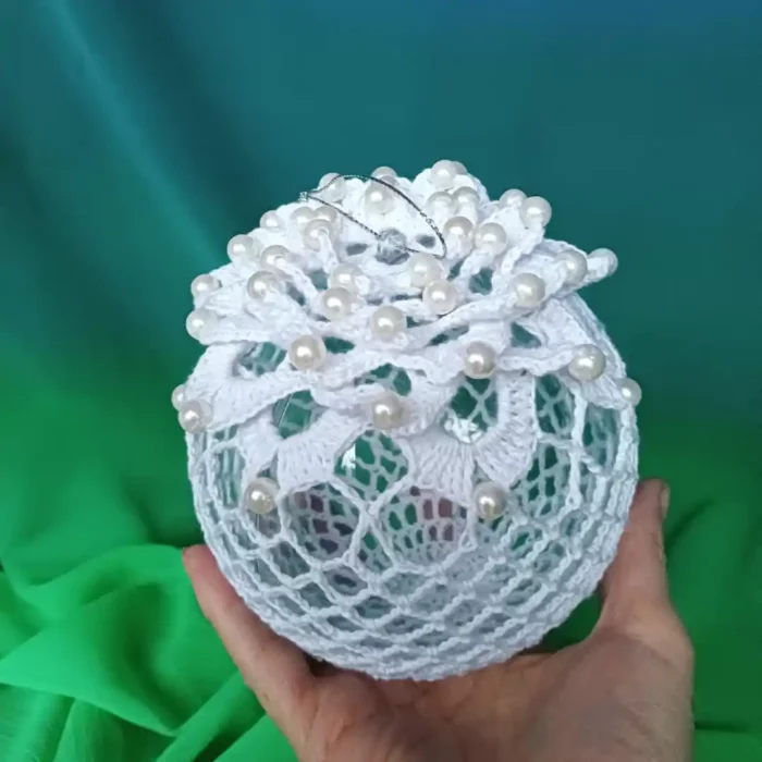 W dłoni bombka szydełkowa zrobiona z białego kordonka wzorem z kwiatem ozdobionym koralikami w kolorze perłowym. W tle zielony materiał.