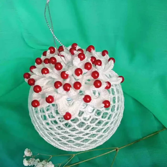 Bombka szydełkowa zrobiona z białego kordonka wzorem z kwiatem ozdobionym koralikami w kolorze perłowym. W tle zielony materiał.