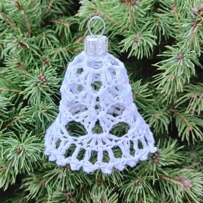 Mały dzwonek zrobiony na szydelku stojący na zielonym iglastym krzewie.