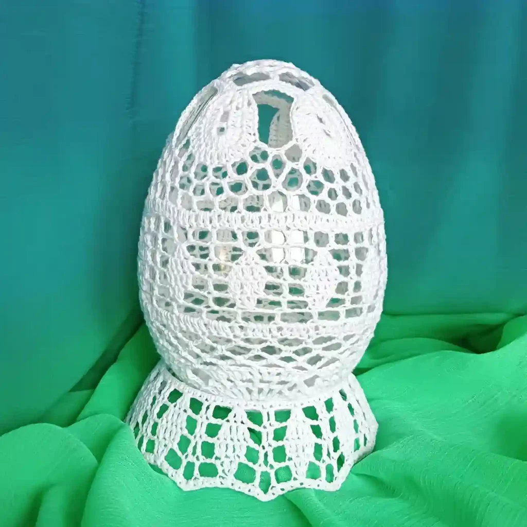 Duże szydełkowe jajko z białego kordonka stojące na podstawce zrobionej na szydełku, w tle zielony materiał
