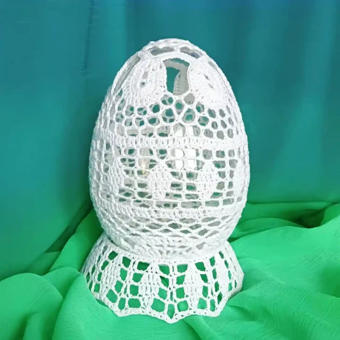Duże szydełkowe jajko z białego kordonka stojące na podstawce zrobionej na szydełku, w tle zielony materiał