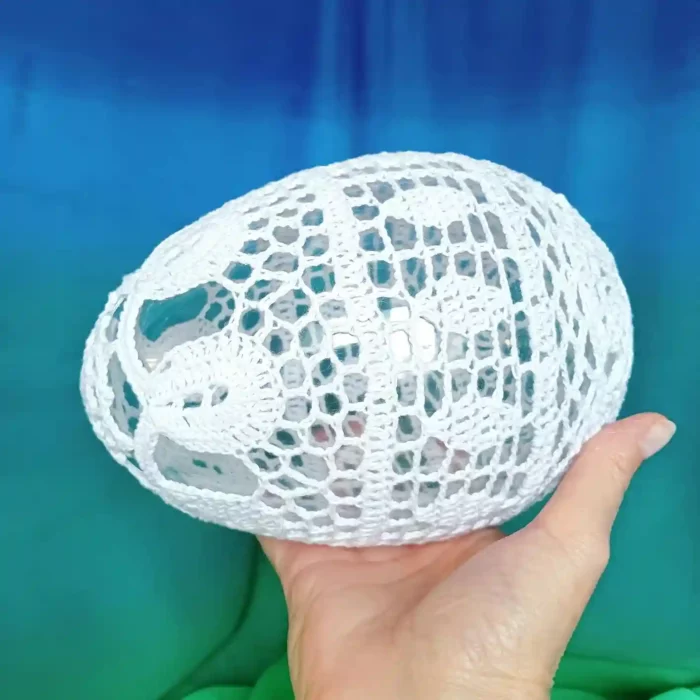 W wyciągniętej dloni duże szydełkowe jajko zrobione na szydelku z bialego kordonka, na wypełnieniu akrylowym