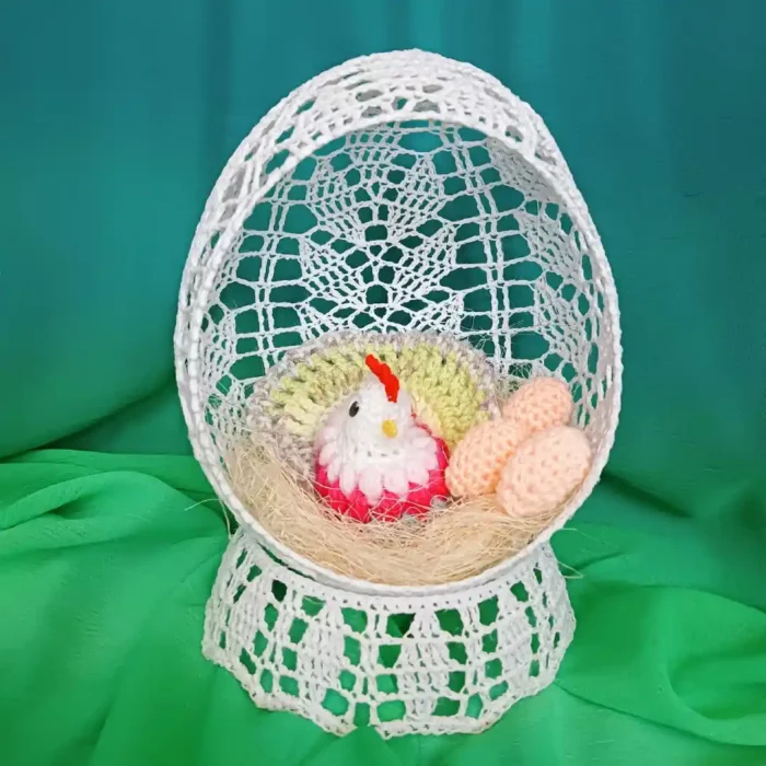 Szydełkowe jajko z dziurka na podstawce, wewnątrz którego znajduje się kolorowa kurka z włóczki siedząca w gniazdku.