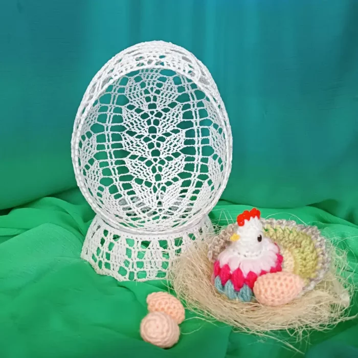 Szydełkowe jajko z dziurka na podstawce obok którego znajduje się kolorowa kurka siedząca w gniazdku i 2 jajeczka zrobione na szydełku.