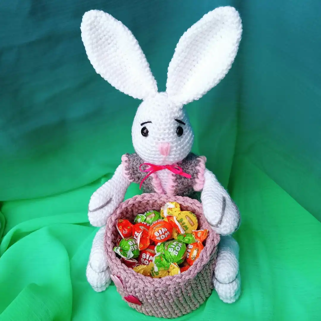 Siedzący królik zrobiony na szydełku z białej włóczki, ubrany w szarą kamizelkę, trzyma koszyczek, w którym są cukierki.