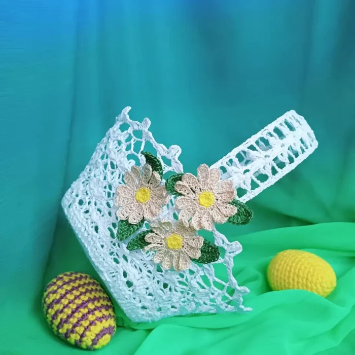 Szydełkowy koszyczek z ozdobnymi stokrotkami zrobiony z białego kordonka na tle zielonego materiału