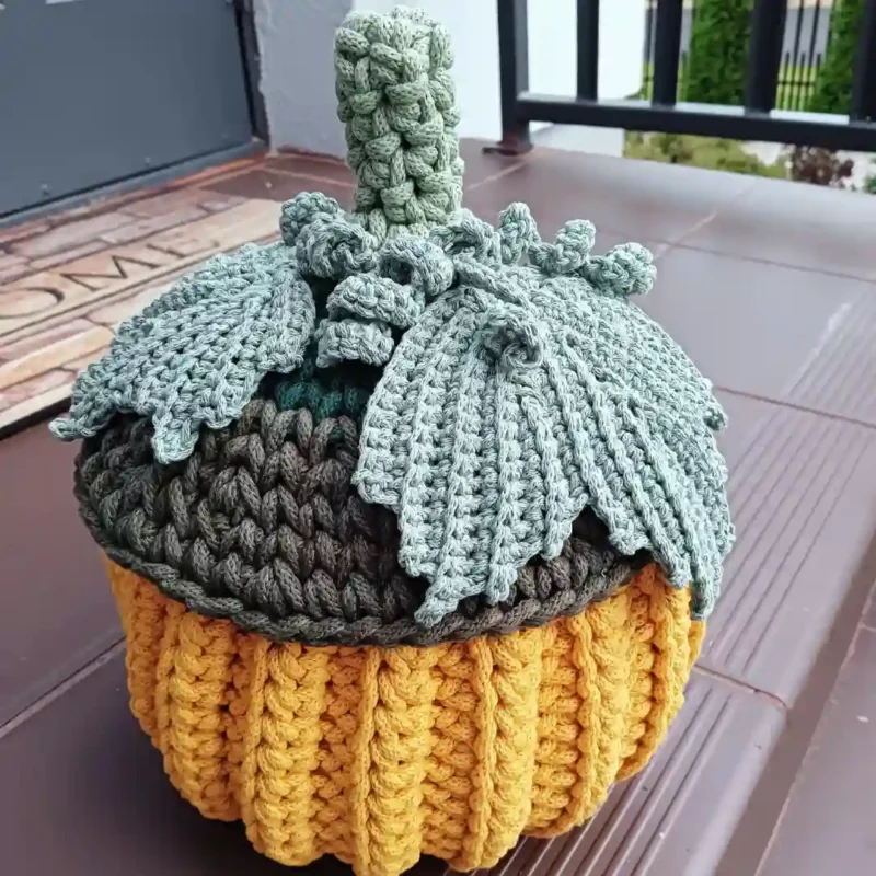 Dynia z przykrywką zrobiona na szydełku ze sznurka bawełnianego, służąca jako pojemnik. Jesienna dekoracja w kolorze żółtym z zieloną przykrywką, ustawiona na schodach przed wejściem.