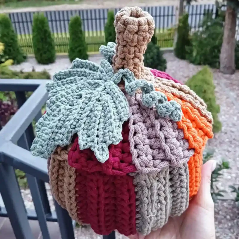 Dynia ze sznurka zrobiona na szydełku_5 Dynia pojemnik na szydełku wykonana z kolorowych bawełnianych sznurków w odcieniach pomarańczowego, beżu, bordo i zieleni, z przykrywką ozdobioną liściem i łodygą, ustawiony na dłoni.