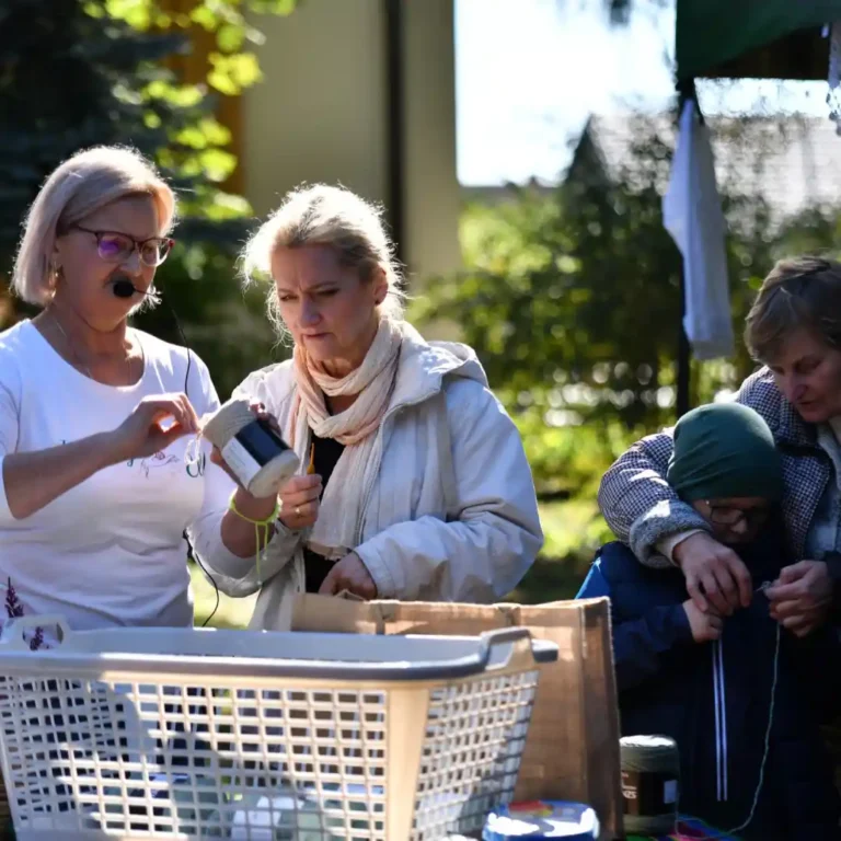 Warsztaty szydełkowania odbywające się na świeżym powietrzu. Instruktorka w białej koszulce i z mikrofonem pokazuje uczestniczce, jak zacząć pracę z włóczką i szydełkiem. W tle inna kobieta pomaga dziecku w zielonej czapce, prowadząc jego ręce podczas nauki.