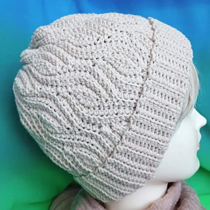 Zdjęcie z prawego profilu jasnobeżowej czapki beanie z tym samym misternym reliefowym wzorem brioszkowym, co na pierwszym zdjęciu. Czapka ma wyraźnie wywinięty brzeg ze ściągaczem. Jest nałożona na głowę manekina, z jasnymi blond włosami wystającymi spod spodu. Manekin nosi pasujący beżowy szalik. Tło jest identyczne: zagięty materiał w odcieniach morskiej zieleni i turkusu.