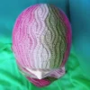Zbliżenie z góry na misternie dzierganą czapkę beanie. Czapka ma trójwymiarowy, pleciony wzór brioszkowy. Przejście tonalne kolorów: od jasnego różu do fioletu po lewej, szaro-beżowa kolumna w środku, zieleń po prawej. Czapka ma wywinięty brzeg, a u dołu widać fragment szyi w pasującym, pasiastym szaliku-kominie. Tło w odcieniach morskiej zieleni i turkusu.