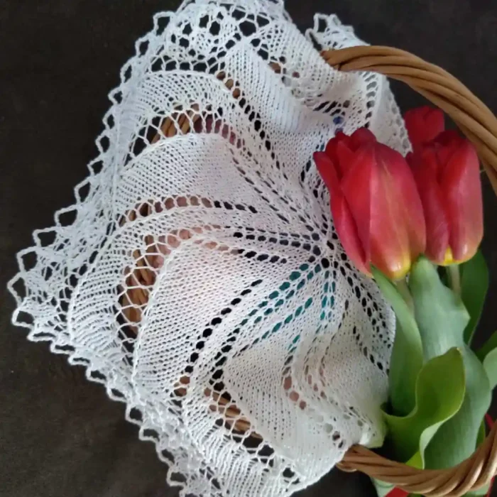 Serwetka-okragla_20 Szczegółowe zbliżenie na pleciony koszyczek z wikliny, w którym ułożona jest koronkowa biała serwetka ręcznie robiona na drutach o charakterystycznym, promieniującym ażurowym wzorze. Serwetka z bawełnianego kordonka częściowo zwisa z koszyczka, a obok niej, w tym samym pojemniku, leży bukiet żywych czerwonych tulipanów ze świeżymi zielonymi liśćmi. Całość sfotografowana na ciemnym tle, tworząc uroczystą, wiosenną i wielkanocną dekorację stołu lub koszyczka.