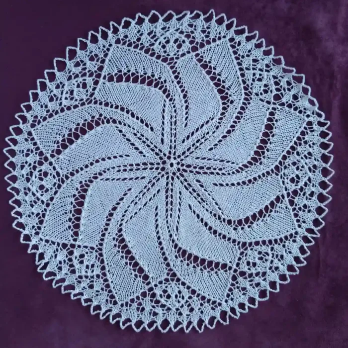 Serwetka-okragla_8 Biała, okrągła serwetka ręcznie robiona na drutach z bawełnianego kordonka. Posiada skomplikowany, mistery ażurowy wzór układający się w promieniującą rozetę lub spiralny wiatraczek, ułożona płasko na ciemnym, purpurowym tle o aksamitnej teksturze.