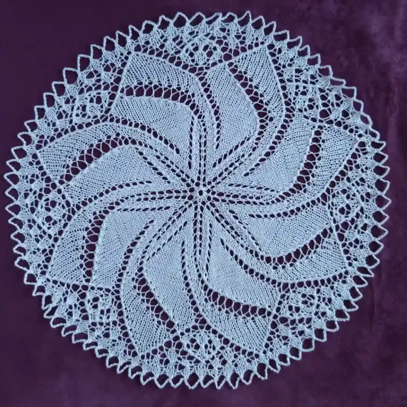 Serwetka-okragla_8 Biała, okrągła serwetka ręcznie robiona na drutach z bawełnianego kordonka. Posiada skomplikowany, mistery ażurowy wzór układający się w promieniującą rozetę lub spiralny wiatraczek, ułożona płasko na ciemnym, purpurowym tle o aksamitnej teksturze.
