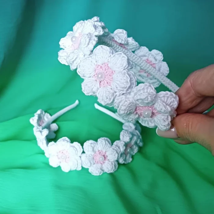 Zbliżenie na precyzyjny, ręczny splot kwiatów: szydełkowa opaska dla dziewczynki z białymi płatkami, różowym środkiem i perłą.