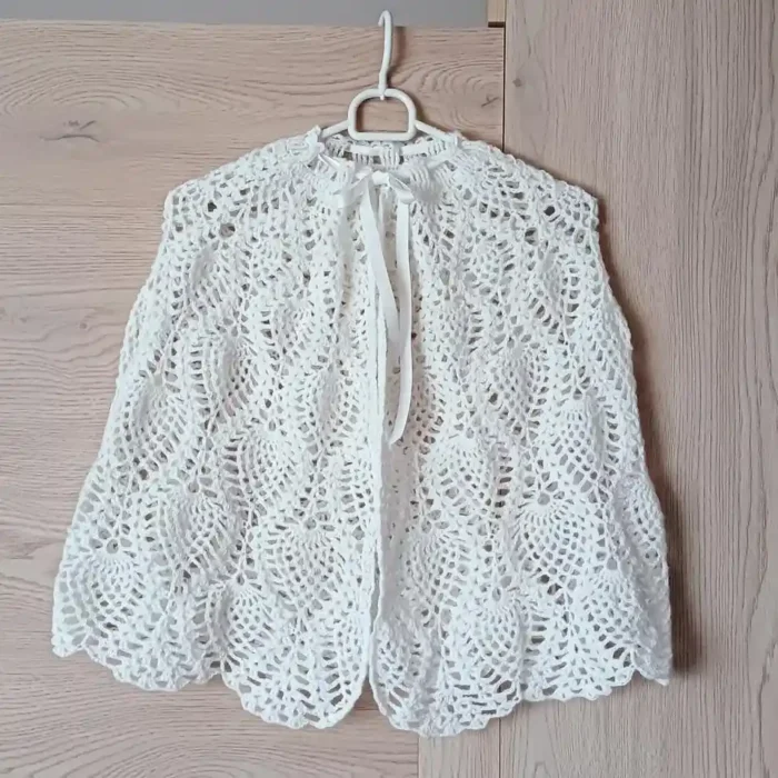 `Pelerynka komunijna - białe szydełkowe bolerko ażurowe z wzorem muszelek, wiązane pod szyją na delikatną satynową wstążkę. Ta elegancka narzutka koronkowa, wisząca na wieszaku na tle drewnianych drzwi, jest idealna na chrzest, pierwszą komunię lub ślub."`