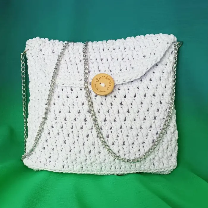 Torebka komunijna ze sznurka poliestrowego w kolorze białym, na srebrnym łańcuszku na tle zielonegomateriału.