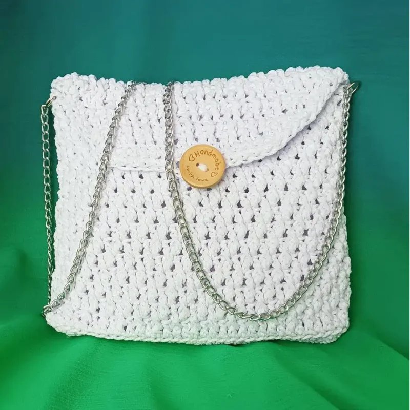 Torebka komunijna ze sznurka poliestrowego w kolorze białym, na srebrnym łańcuszku na tle zielonegomateriału.