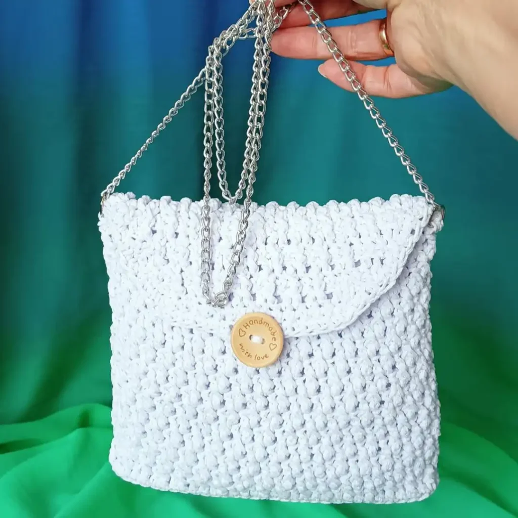 Dłoń trzymająca srebrny łańcuszek białej torebki komunijnej ze sznurka poliestrowego. Torebka ma drewniany guzik z grawerem 'Handmade with love'. Tło z gradientem zielono-niebieskim.