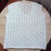 Bezszwowa, biała ażurowa bluzka handmade leżąca na płasko na drewnianym stole, ukazująca z bliska misterny, geometryczny wzór z włóczki.