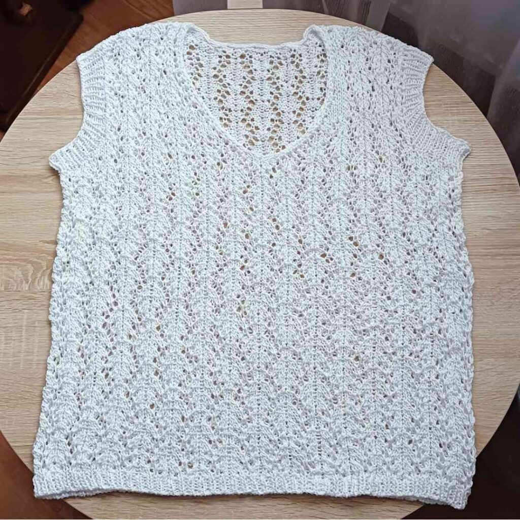 Bezszwowa, biała ażurowa bluzka handmade leżąca na płasko na drewnianym stole, ukazująca z bliska misterny, geometryczny wzór z włóczki.