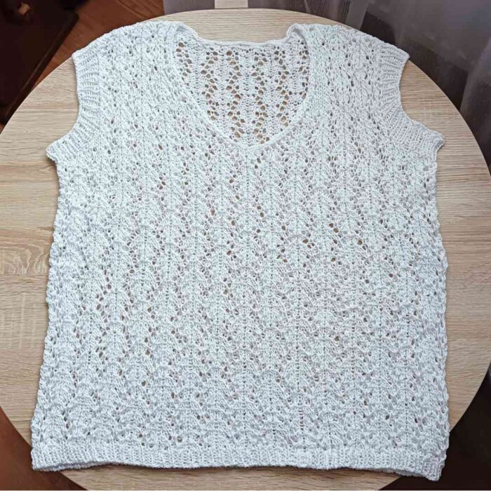 Bezszwowa, biała ażurowa bluzka handmade leżąca na płasko na drewnianym stole, ukazująca z bliska misterny, geometryczny wzór z włóczki.