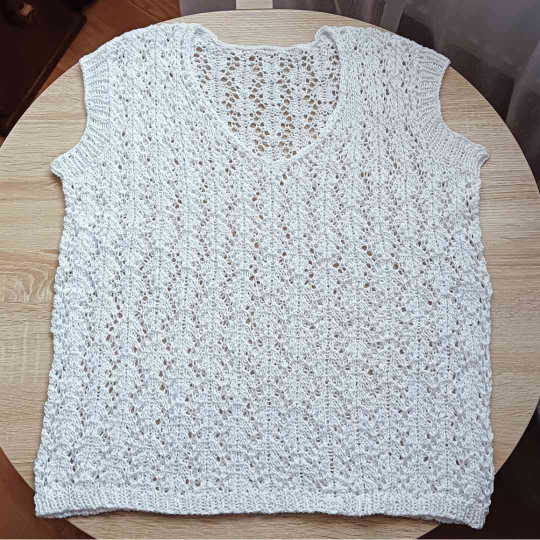Bezszwowa, biała ażurowa bluzka handmade leżąca na płasko na drewnianym stole, ukazująca z bliska misterny, geometryczny wzór z włóczki.