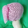 Chustka bandana zrobiona na szydełku_7 Tył głowy manekina w różowej szydełkowej chustce z węzłem, na zielonym tle.