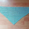 Rozłożona na płasko trójkątna, jasnobłękitna, szydełkowana chustka bandana z ażurowym wzorem, leżąca na drewnianym blacie stołu.