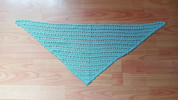 Rozłożona na płasko trójkątna, jasnobłękitna, szydełkowana chustka bandana z ażurowym wzorem, leżąca na drewnianym blacie stołu.