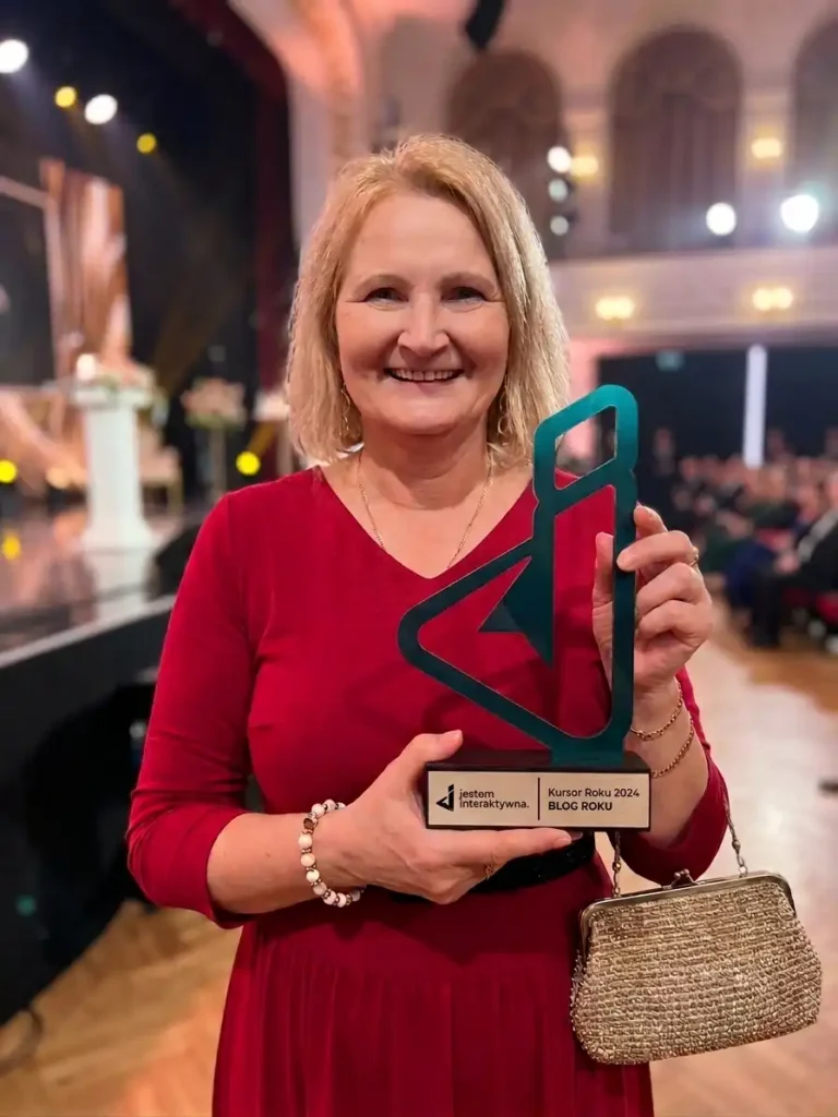 Uśmiechnięta Elżbieta (właścicielka Bajery Eli) w czerwonej sukience stoi na scenie podczas gali, trzymając w rękach nowoczesną, turkusową statuetkę nagrody „Kursor Roku 2024” w kategorii „BLOG ROKU”. W tle widać rozmytą widownię i scenografię wydarzenia.