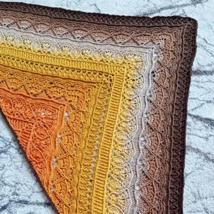 Zbliżenie na misterną fakturę i geometryczny, ażurowy wzór ręcznie robionej szydełkowej chusty w odcieniach miodowego żółtego, beżu i ciepłego brązu